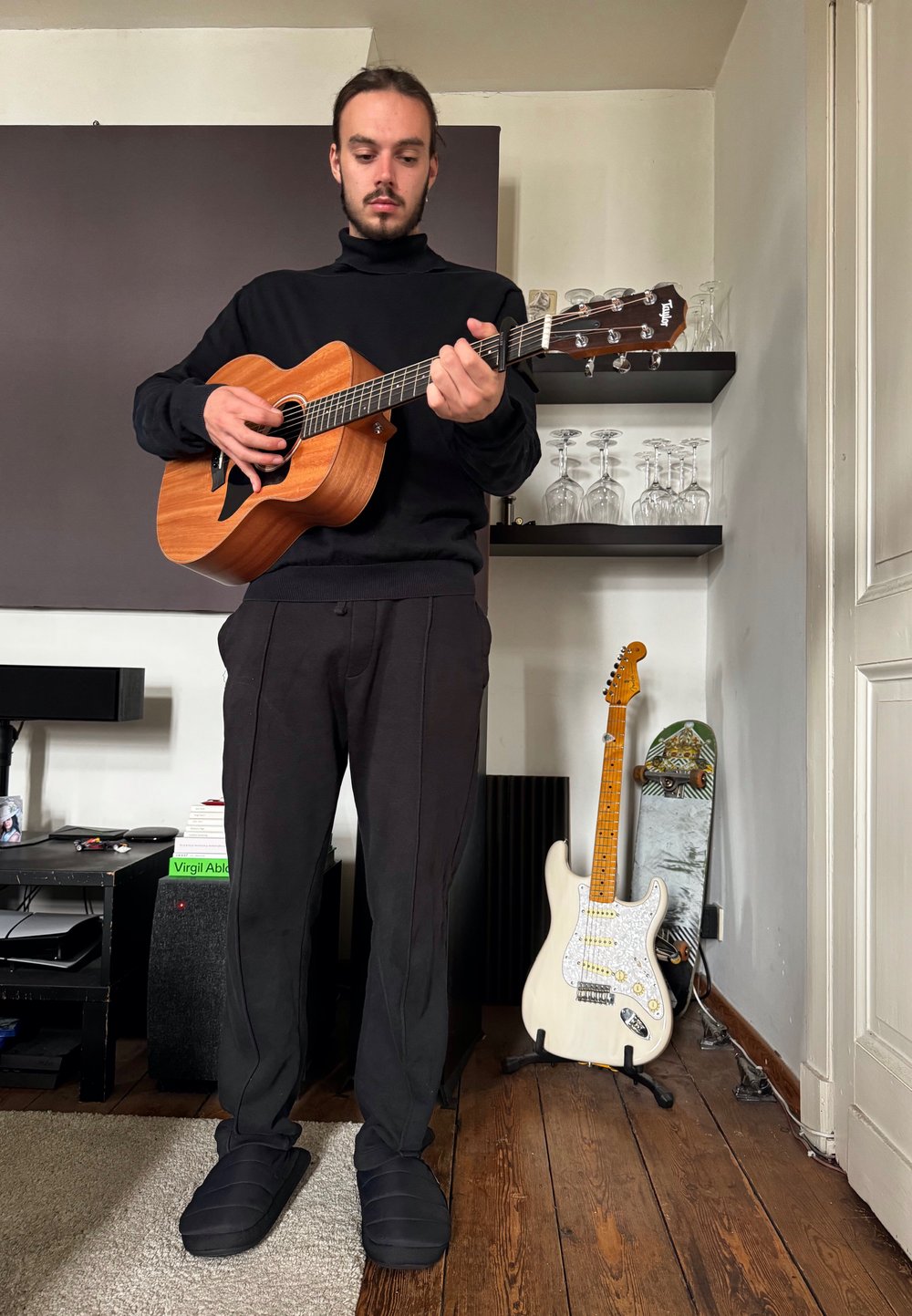 Homem segurando uma guitarra acústica de madeira, vestindo um suéter preto de gola alta e calças de moletom. Chinelos pretos, chão de madeira clara e prateleiras com vidro atrás.