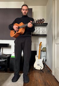 Homem segurando uma guitarra acústica de madeira, vestindo um suéter preto de gola alta e calças de moletom. Chinelos pretos, chão de madeira clara e prateleiras com vidro atrás.