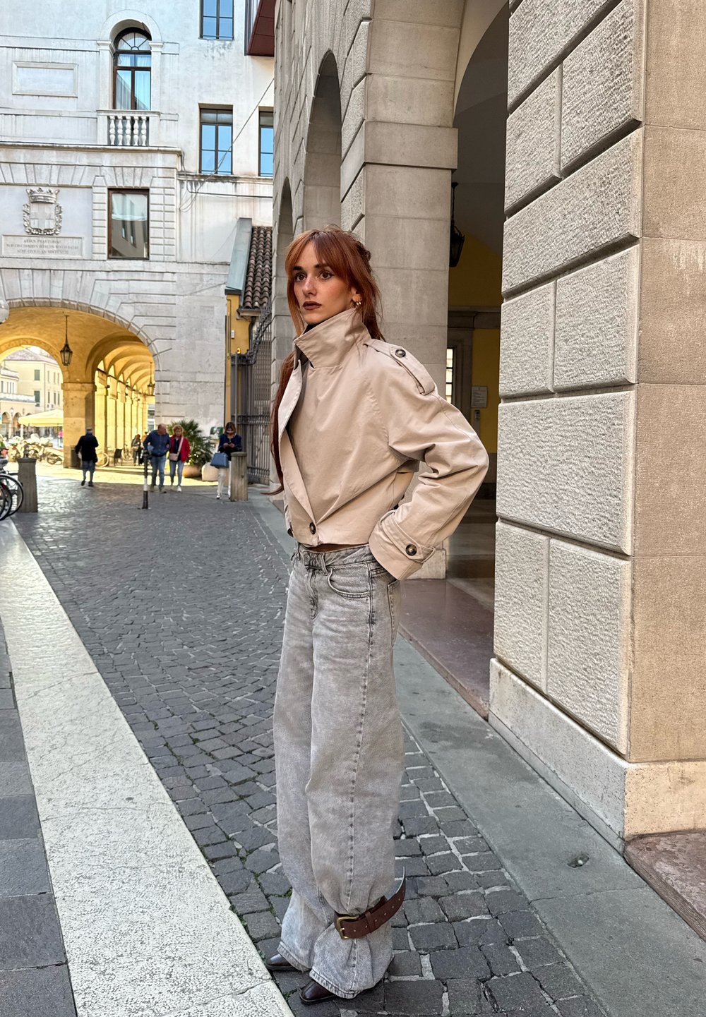 Beige zkrácená bunda s vysokým límcem, doplněná světle šedými kalhotami s širokými nohavicemi. Tmavý pásek v pase. Hladké textury a jemné barvy v městském prostředí.