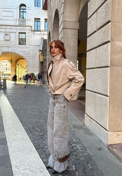 Chaqueta beige corta con cuello alto, combinada con jeans de pierna ancha en gris claro. Cinturón oscuro en la cintura. Texturas suaves y colores delicados en un entorno urbano.