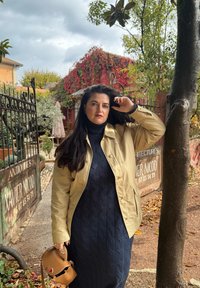 Lys kremfarget jakke over en mørk marineblå strikkekjole, holdt med den ene hånden. En liten brun håndveske henger på siden, med en teksturert finish.