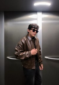 Jeune homme portant des lunettes de soleil noires et une veste en cuir marron, debout à l'intérieur d'un ascenseur métallique sous une lumière intense au plafond.