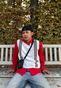 Άτομο που φοράει ένα λευκό πουλόβερ με μαύρες ρίγες πάνω από ένα κόκκινο πουκάμισο, μαύρο καπέλο και μπλε τζιν, καθισμένο σε ένα παγκάκι με πράσινα φυτά από πίσω.