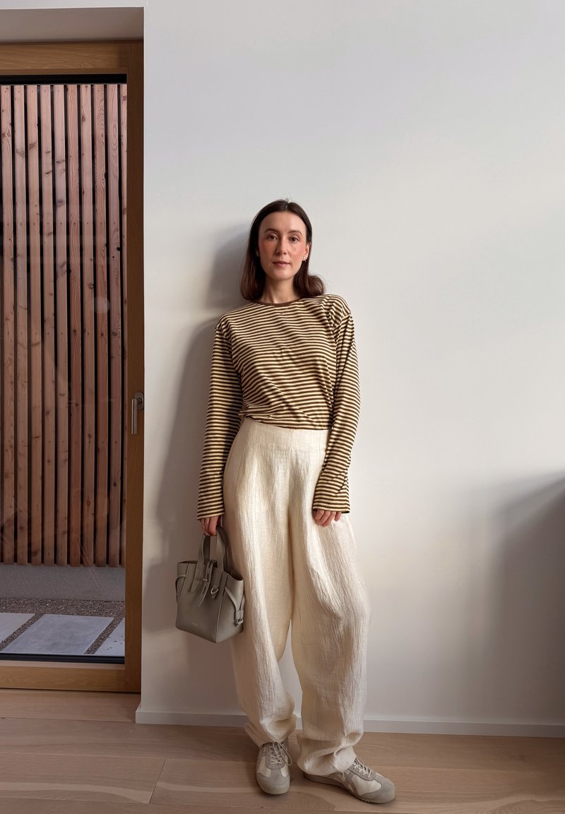 Femme aux cheveux foncés portant un haut à manches longues beige et marron à rayures, pantalon ample crème, baskets, tenant un sac à main gris, debout à l'intérieur.