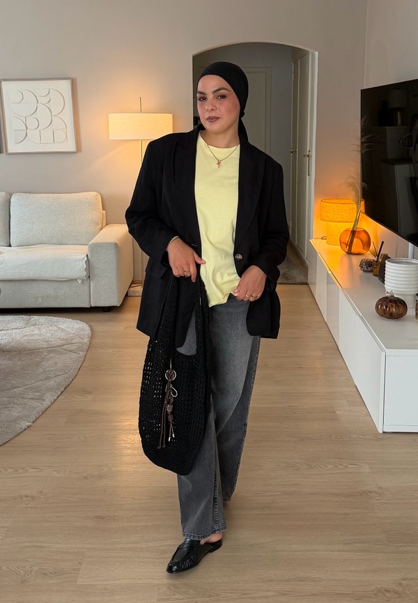 Femme portant un foulard noir, un blazer noir, une chemise jaune, un jean gris, tenant un sac tricoté noir, debout dans un salon moderne.