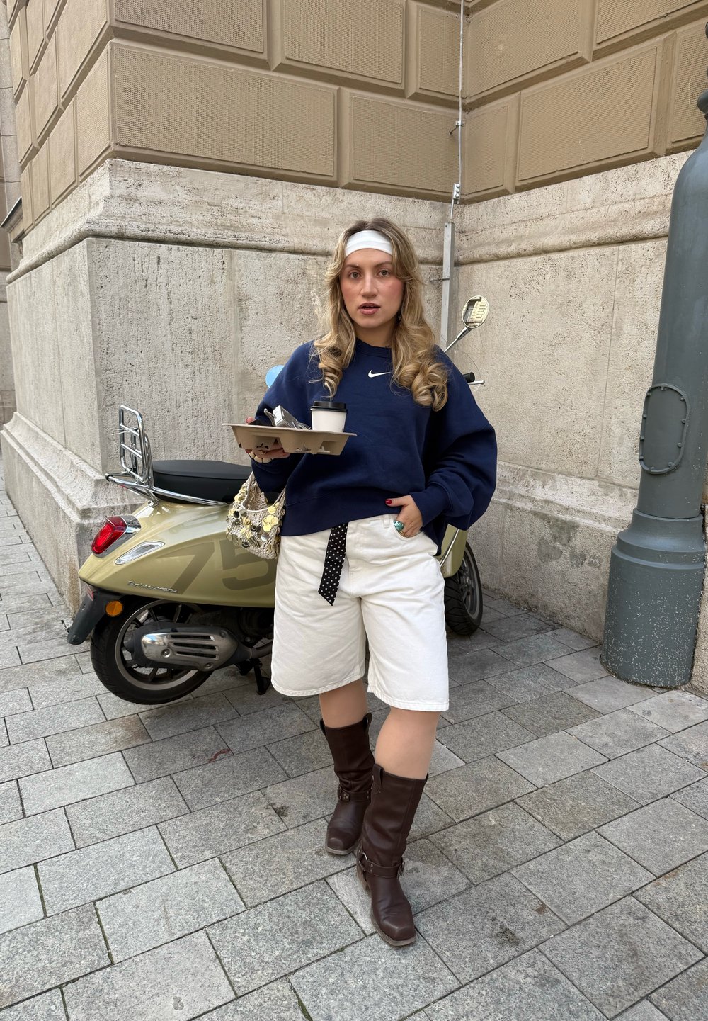 Felpa blu navy, pantaloni corti bianchi, stivali marroni fino al ginocchio. Tiene una tazza di caffè e un vassoio con posate. Scooters sullo sfondo.