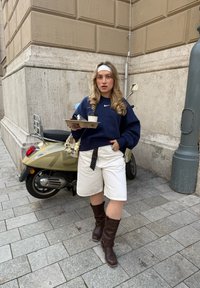 Felpa blu navy, pantaloni corti bianchi, stivali marroni fino al ginocchio. Tiene una tazza di caffè e un vassoio con posate. Scooters sullo sfondo.