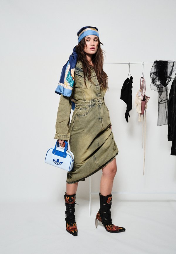 Femme en robe verte avec foulard bleu à motifs tient un sac Adidas bleu, porte des bottes noires à motif de flammes, debout près d'un porte-vêtements avec des vêtements suspendus.