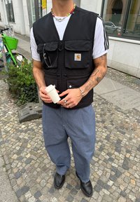Črna zip-up brezrokavnik z dvema sprednjima žepoma, nošen čez belo majico. Modri široki hlače in črne čevlje. Drži bel skodelico.