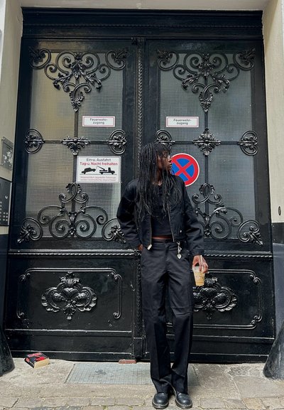 Chaqueta negra y pantalones usados por una persona de cabello largo, de pie junto a unas puertas dobles negras ornamentadas con acentos de metal y letreros decorativos.