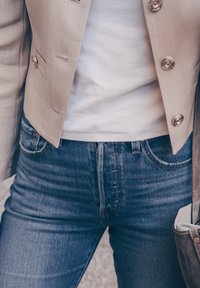 Nærbilde av en person som har på seg blå jeans, hvit skjorte, beige jakke med knapper, og bærer en grå suedepose over skulderen.