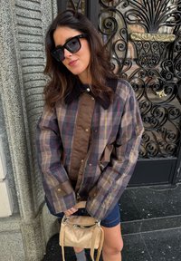 Brūnas un pelēkas šaha raksta jakas ar korduroja apkakli, kopā ar mazu bēša krāsas somu. Tumši džinsa šorti un saulesbrilles noslēdz šo izskatu.