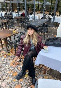 Mørkerød læderjakke, grå sweater, sorte blondebukser, sorte støvler og béret. Holder en kaffekop ved et bord omgivet af efterårsblade.