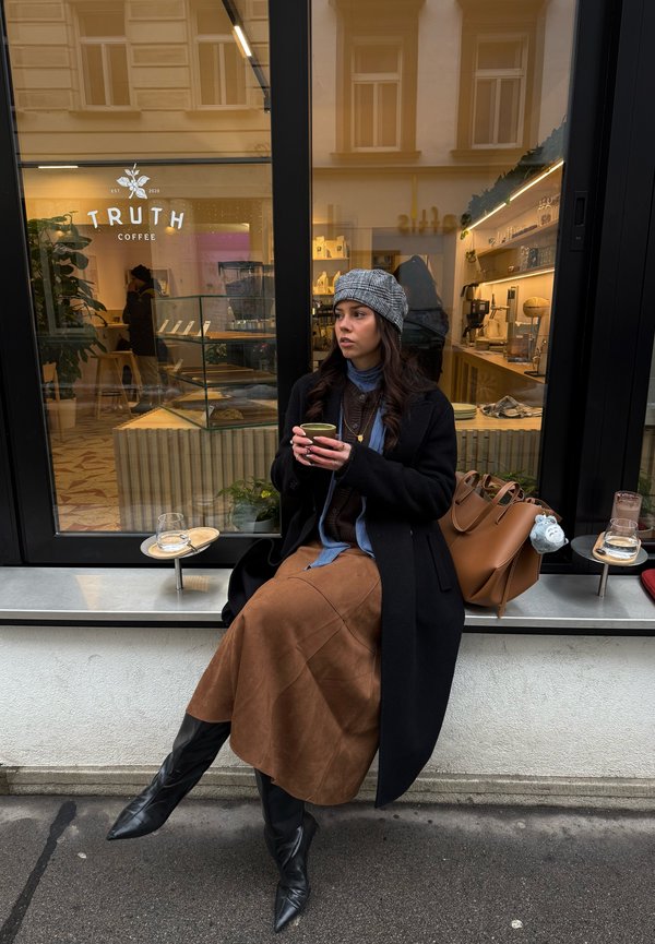 Femme portant un chapeau gris et un manteau noir, assise devant un café, tenant une tasse verte, avec un sac marron à côté d'elle.