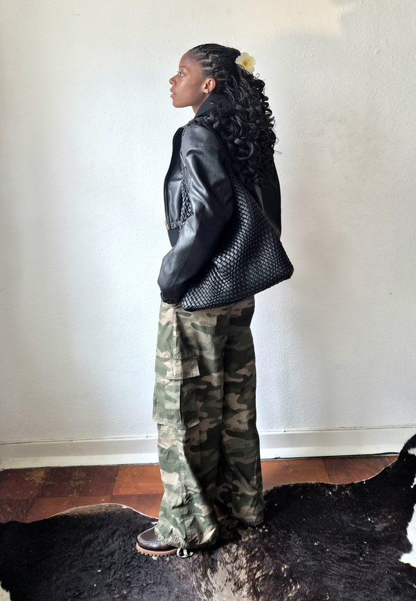 Joven con trenzas y accesorio de flores lleva chaqueta de cuero negra, gran bolso tejido negro, pantalones de camuflaje y botas marrones, de pie en el interior.