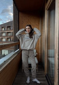 Sweat à capuche surdimensionné beige clair avec le texte "LAMANIA" et un pantalon de jogging assorti. Baskets blanches avec des rayures noires. Balcon en bois et en verre.