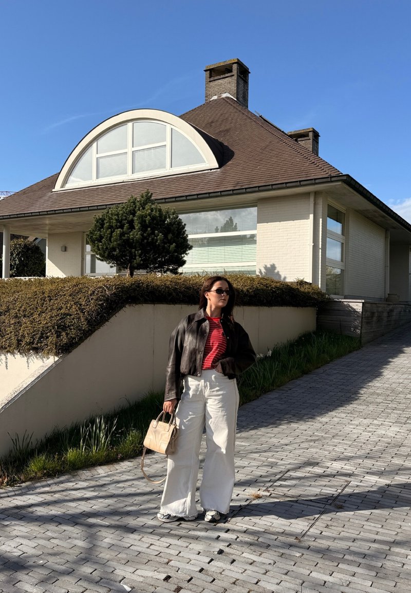 Woman wearing sunglasses, black leather jacket, red striped shirt, white wide-leg pants, holding beige handbag, standing on paved path by white house.