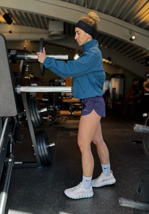Mujer con ropa deportiva ajustando placas de peso en una máquina de gimnasio en un centro de fitness con equipo al fondo.