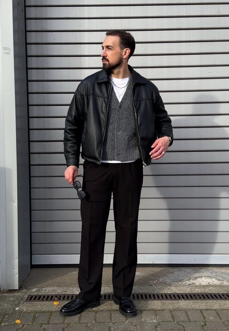 Giacca di pelle nera sopra un cardigan grigio, t-shirt bianca e pantaloni neri. Tiene occhiali da sole, indossa mocassini neri. Sfondo a righe.