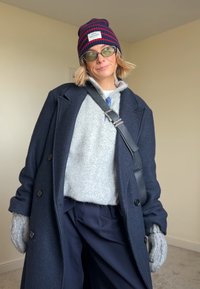 Manteau en laine bleu marine, pull gris duveteux, pantalon large bleu marine, gants en tricot gris et bonnet rayé avec une étiquette.