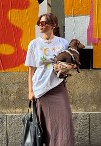 T-shirt gráfica branca com impressão colorida, saia maxi castanha, bolsa tote preta, óculos de sol, segurando um cão castanho com um arnês, em frente a uma parede colorida.