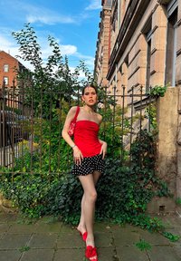 Top rosso senza spalline abbinato a una gonna nera a pois, sandali rossi e una borsa a fantasia in un ambiente verde all'aperto.