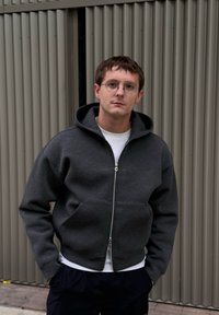 Sweatshirt cinza com fecho de correr e capuz, com bolso frontal e forro interior branco, usado por cima de uma t-shirt branca. Tecido texturizado, design casual.