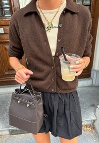 Cardigan castanho sobre uma blusa canelada bege, saia pregueada preta. Segura uma mala estruturada castanho-escura e um copo transparente com um palhinha.