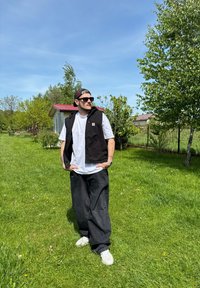 Schwarze ärmellose Weste über einem weißen T-Shirt, kombiniert mit schwarzen lockeren Hosen und weißen Sneakers. Die Szene zeigt grünes Gras und Bäume.