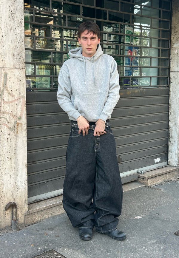 Junge Person in übergroßen, weiten Jeans und grauer Kapuzenjacke steht auf städtischem Bürgersteig vor einem Metallrollladen.