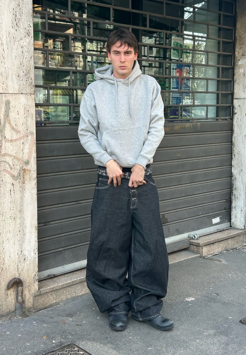 Mladý človek v nadrozmerných voľných džínsach a šedej mikine s kapucňou stojí na mestskom chodníku pri kovovej závore.