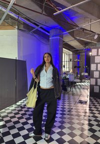 Eine Person steht in einem geräumigen Raum, trägt ein hellblaues, kurzes Shirt, schwarze weite Hosen und silberne Sneakers und hält eine gelbe Tote-Tasche.