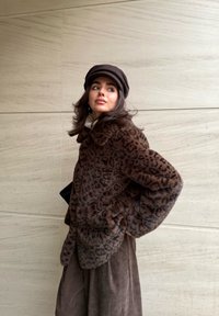 Veste en fausse fourrure marron à motif léopard, manches longues et col arrondi, assortie d'une casquette foncée et d'un pull beige en dessous.