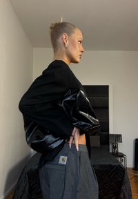 Femme aux cheveux blonds en chignon, portant une veste noire et un pantalon gris, debout de profil dans une chambre avec un lit et une lampe en arrière-plan.