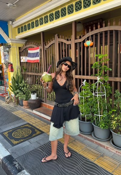 Mujer con sombrero negro, gafas de sol y ropa en capas, sosteniendo un coco con una pajilla, de pie en una acera urbana con patrones cerca de macetas.