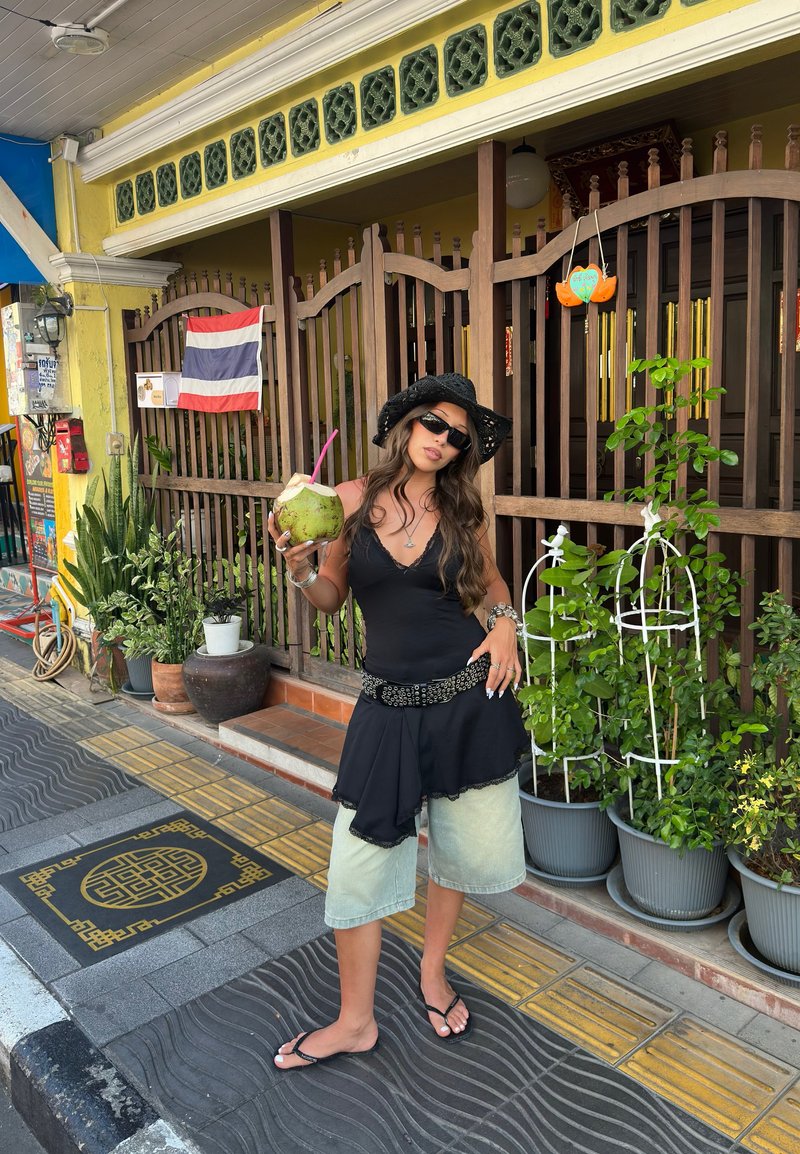 Γυναίκα με μαύρο καπέλο, γυαλιά ηλίου και στρωμένη ένδυση κρατάει καρύδα με καλαμάκι, στέκεται σε πεζοδρόμιο με σχέδια κοντά σε γλάστρες.