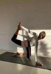 Træningsudstyr i lys grå og mørkebrun, tekstureret stof. Person udfører en yogaøvelse på en mørk måtte, mod en lys væg.