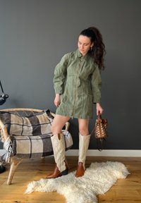 Robe veste vert olive avec fermeture éclair, poches latérales et manches longues. Bottes de cowgirl crème avec accents marron. Sac seau marron clouté.