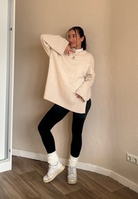 Pull beige clair oversized avec ourlet côtelé, porté par-dessus des leggings noirs. Les accessoires incluent des baskets blanches et des jambières blanches confortables.