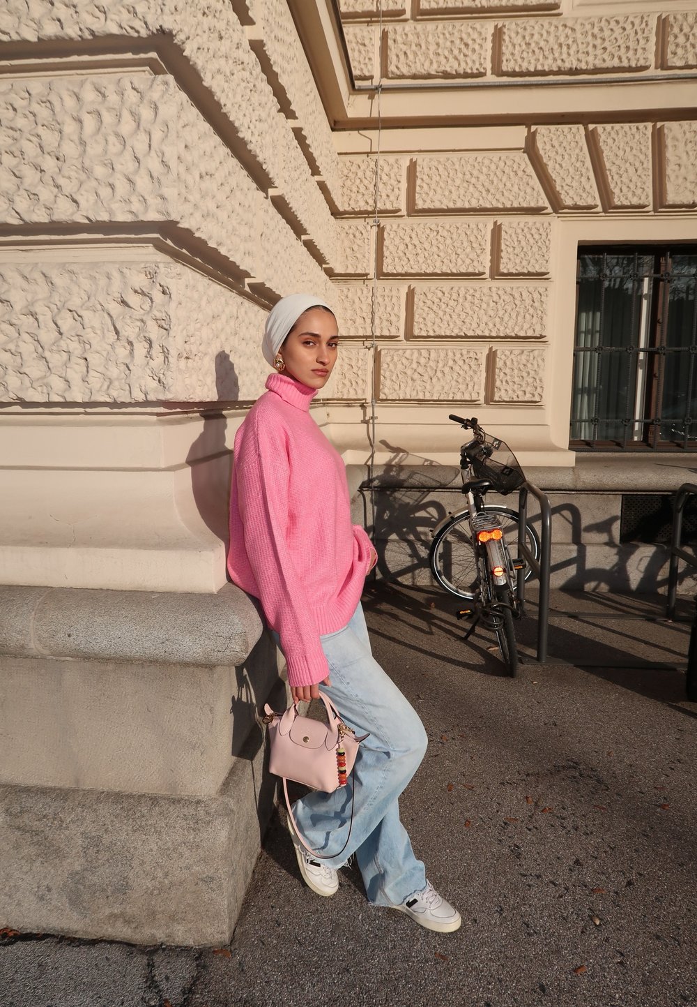 Pullover rosa lavorato a maglia, jeans a gamba larga blu chiaro, tiene una piccola borsa rosa con una tracolla decorativa. Parete testurizzata e bicicletta sullo sfondo.