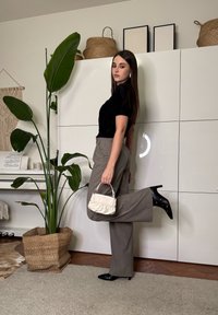 Γυναίκα με μακριά σκούρα μαλλιά ποζάρει μέσα σε εσωτερικό χώρο, φοράει μαύρο τοπ, γκρι παντελόνι με φαρδιά μπατζάκια, μαύρα μποτάκια με τακούνι και κρατά μικρή λευκή τσάντα χειρός.