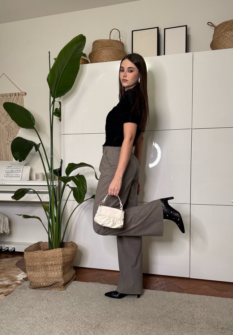 Frau mit langen dunklen Haaren posiert drinnen, trägt ein schwarzes Oberteil, graue Schlaghosen, schwarze Stiefel mit Absatz und hält eine kleine weiße Handtasche.