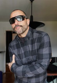 Camicia di flanella a quadri neri e grigi, abbinata a occhiali da sole e un orecchino distintivo. Ambiente interno con elementi di arredamento a contrasto.