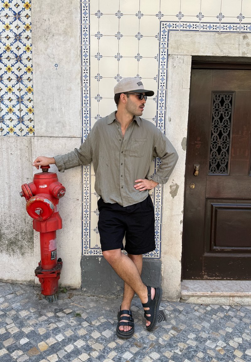 Homme portant des lunettes de soleil, une casquette, une chemise verte et un short noir appuyé sur une bouche d'incendie rouge à côté d'un mur carrelé et d'une porte en bois sombre dans une rue pavée.