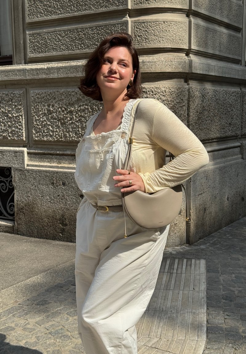 Junge Frau in cremefarbenen Overalls und langärmeligem Shirt posiert mit einer beigefarbenen Handtasche an einer Kopfsteinpflasterstraße in der Sonne.