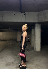 Top din piele neagră și pantaloni negri conturați, sandale cu platformă groasă negre, ținând o geantă de mână roz cu un model, cu un fundal subteran.