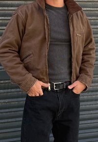 Mann trägt eine braune Jacke mit Reißverschluss, ein dunkles, geripptes Hemd, schwarzen Gürtel und schwarze Hose, hat die Hände in den Taschen und steht vor einem Hintergrund aus wellblechmetal.