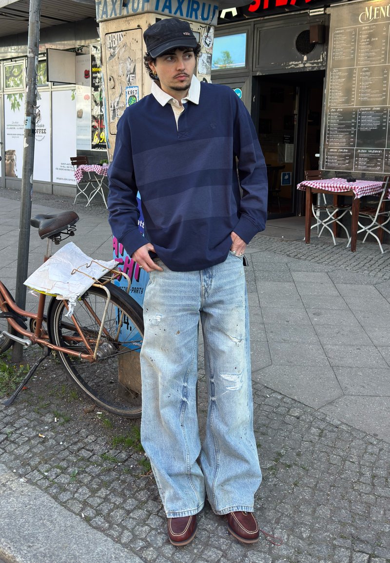 Jeune homme portant un pull bleu marine, un jean délavé à jambes larges et des chaussures marron, debout sur un trottoir en ville à proximité d'un vélo et d'un café.