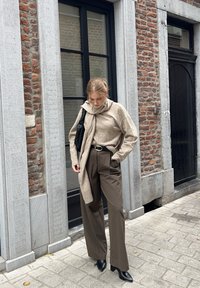 Frau in beigem Pullover und Schal, braunen weiten Hosen, schwarzen Stiefeln und mit einer schwarzen Schultertasche, die auf einer Kopfsteinpflasterstraße steht.