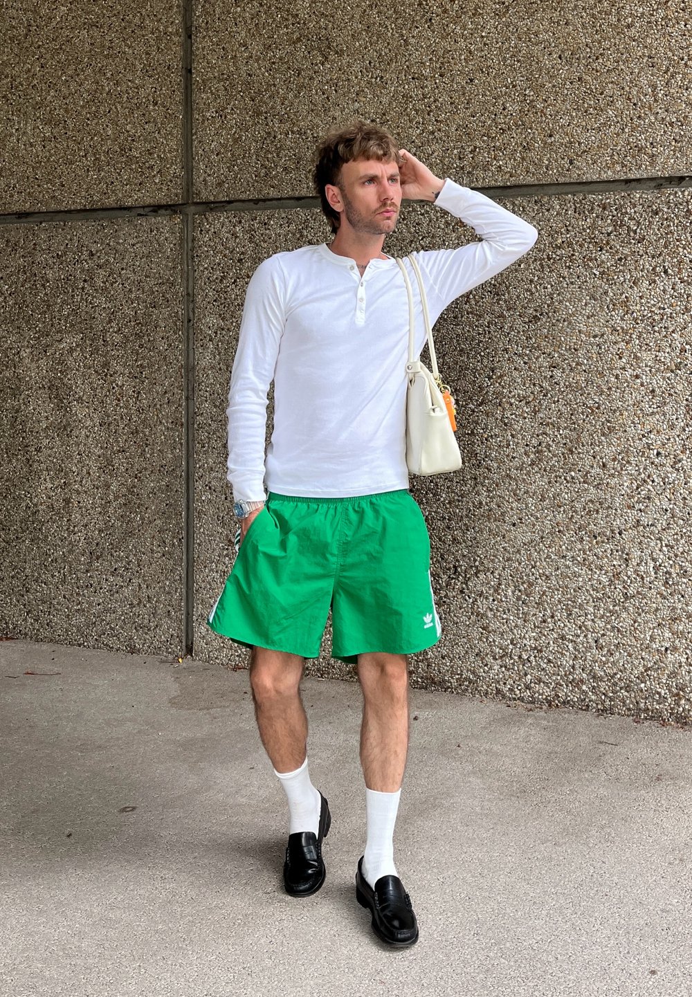 Weißes Langarmhemd, grüne Shorts, schwarze Slipper und weiße Socken. Bemerkenswerte Details sind ein kleines Markenlogo und eine weiße Handtasche.